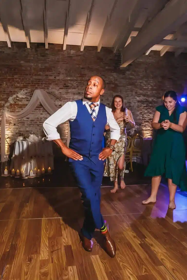 Man in a blue three-piece suit dancing confidently on a wooden floor at a party, with two smiling women watching in the background against a brick wall backdrop.
