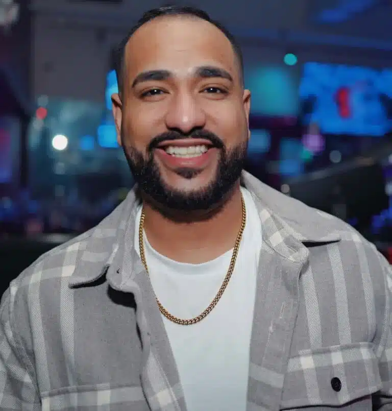 Smiling man with a beard in a white T-shirt and gray plaid shirt, wearing a gold chain, in a dim nightclub setting.