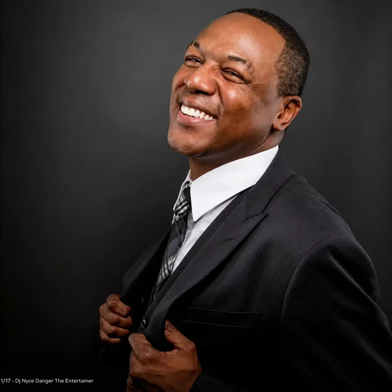 Smiling man in a dark suit adjusting his jacket against a black background.