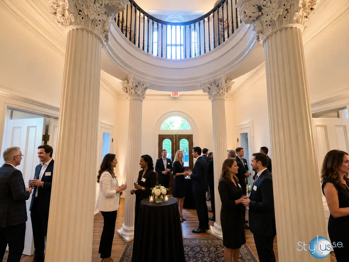 Formal networking event in a grand columned foyer with people chatting around a tall round table in the center.