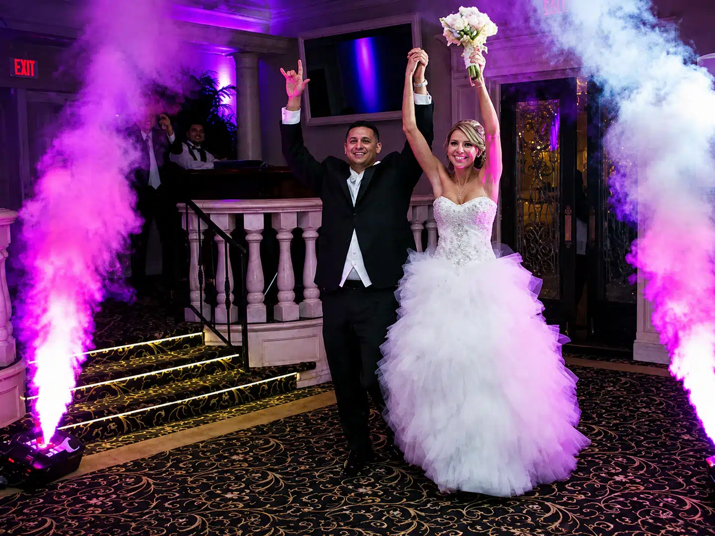 Bride in a white wedding gown and groom in a black tuxedo raise their hands in celebration as purple smoke surrounds them at a wedding venue in a ballroom setting, bouquet raised by the bride.
