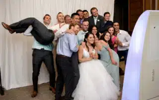 Group of wedding guests posing for a photo, bride in a white gown front center, friends lift a man on the left, all smiling in formal attire in front of a white backdrop and a photo booth.