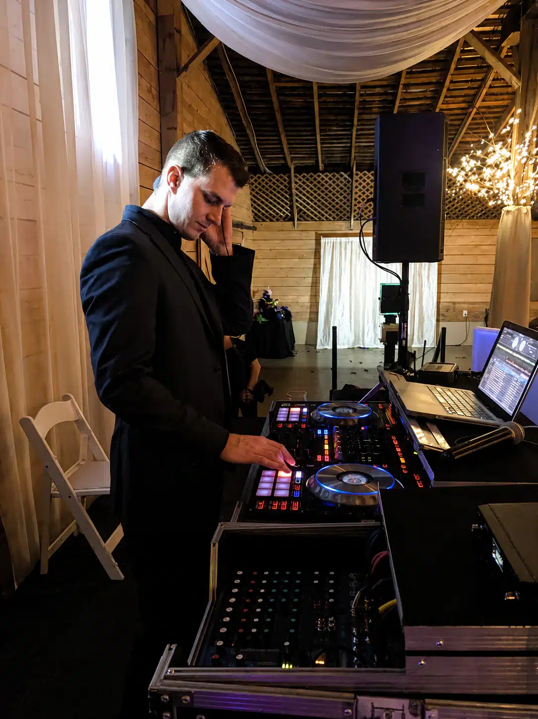 DJ in a black suit operating a lit DJ controller with a laptop nearby at a wooden-venue event space