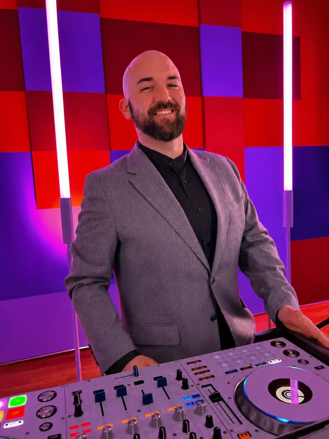 Smiling man in a gray suit behind a DJ mixer in a colorful studio with red and purple wall panels and vertical neon lights.