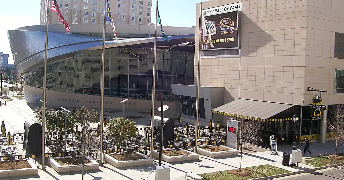 Exterior view of the NASCAR Hall of Fame building with a curved glass front, flagpoles, and an outdoor dining area in front of Buffalo Wild Wings.