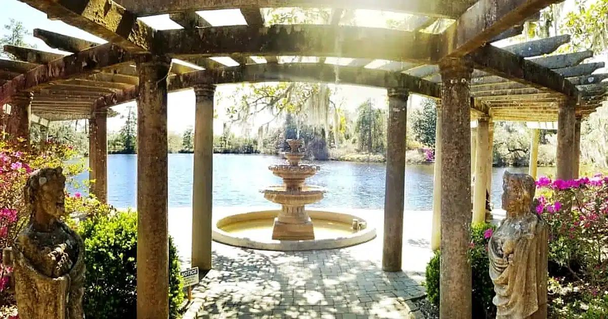 Pergola-covered courtyard with a tiered fountain by a lakeside, flanked by stone statues and blooming shrubs.
