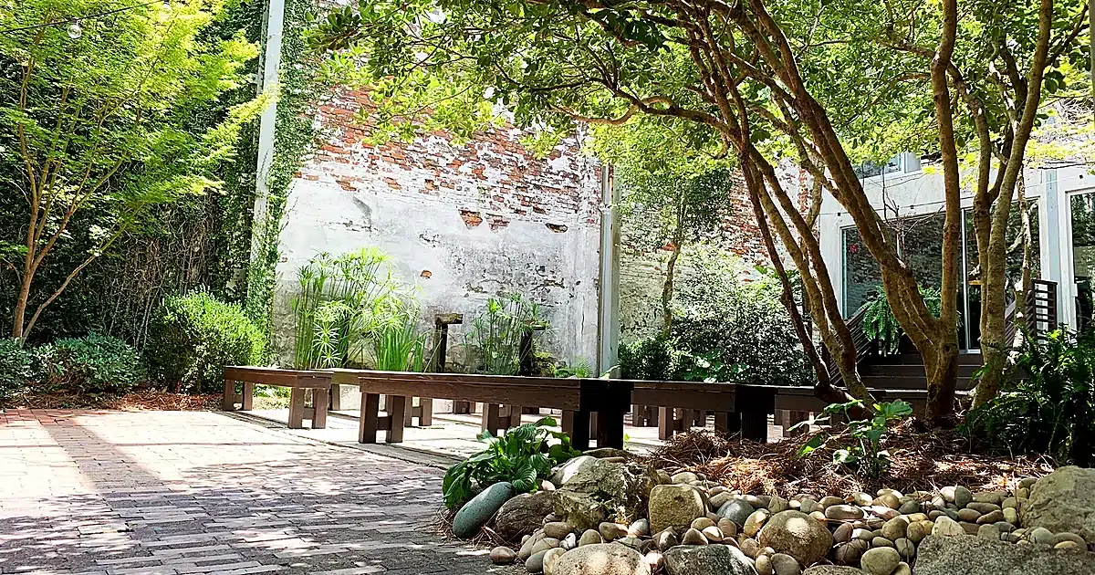 Courtyard with brick wall, wooden benches, and a pebble garden under leafy trees.
