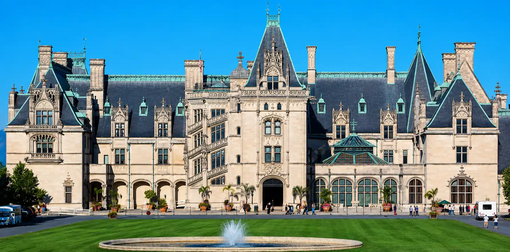 Gothic-style university building with ornate stonework, slate roof, and towers, facing a circular fountain and green lawn.