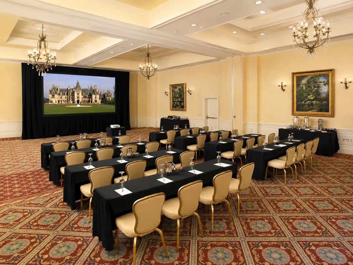 Elegant conference room set up for a meeting with black-clothed tables, beige chairs, chandeliers, and a large screen showing a castle.