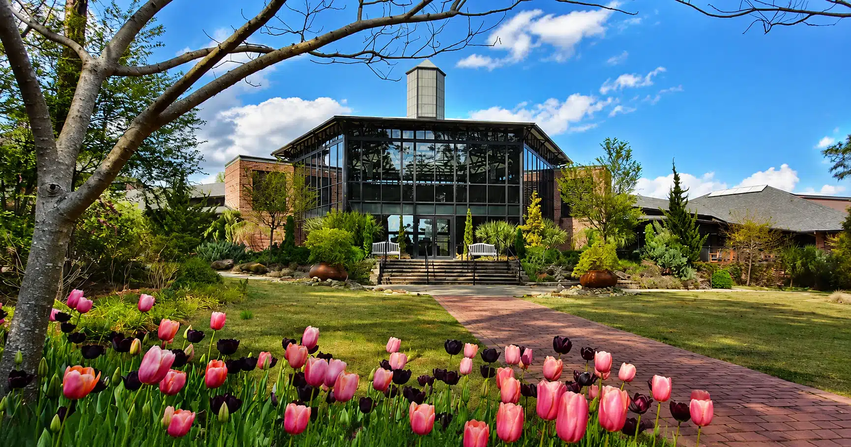 Brick-and-glass conservatory-style building with a glass atrium, surrounded by a manicured garden and tulips.