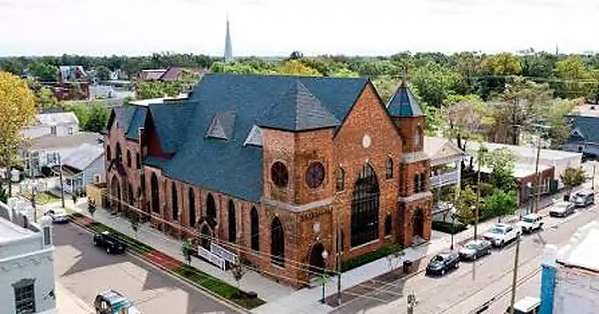 A brick church with a blue slate roof and tall steeple on a street corner in a small town.