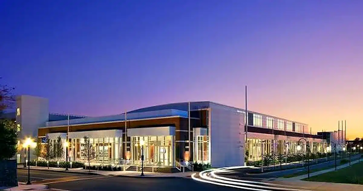 Modern glass-front commercial building at dusk with warm interior lights and a curved driveway in front.