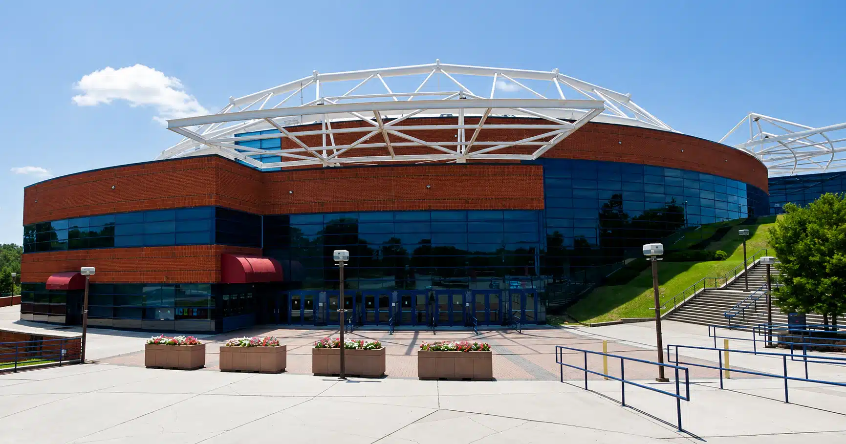 Modern brick and glass arena with a white steel lattice roof against a blue sky, blue-tinted windows and entry plaza.