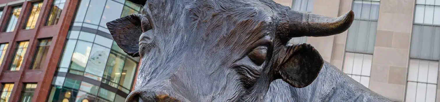 Bronze bull statue in an urban plaza with glass-fronted buildings behind it.