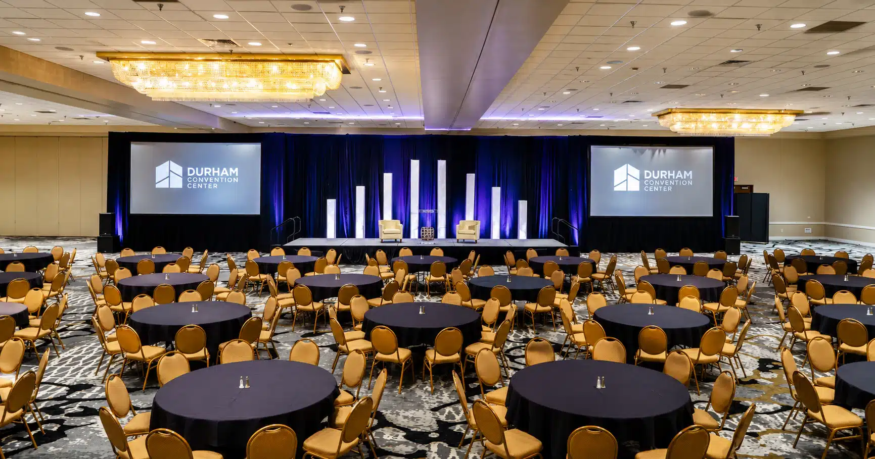 Conference hall with round tables and gold chairs facing a stage with blue curtains and two screens showing Durham Convention Center logo.