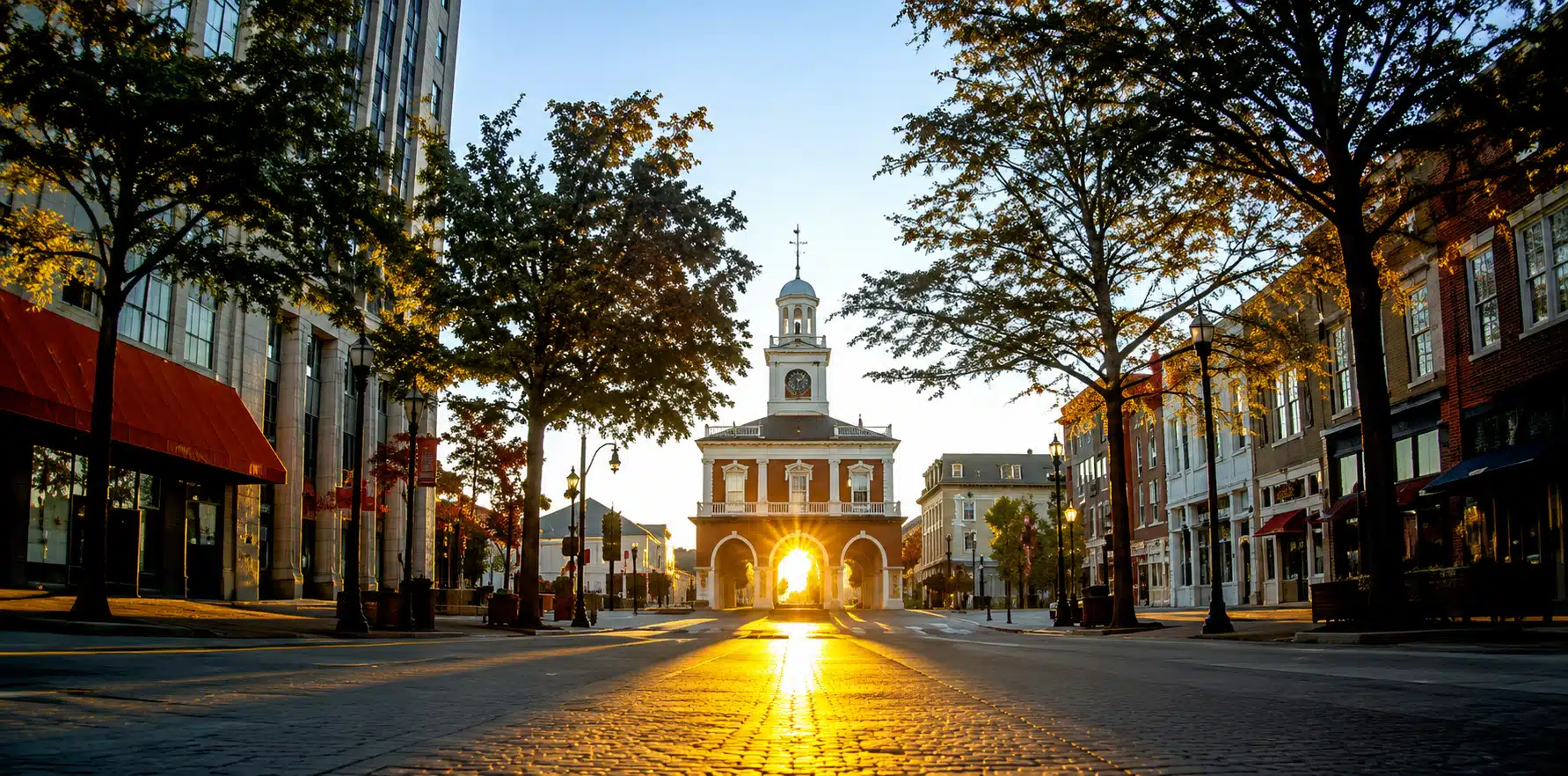 Sunlit street toward a central clock tower arch building, with golden glow streaming through the arches at sunset.