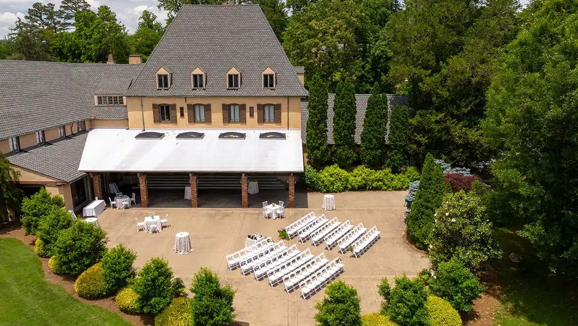 Outdoor event setup in a courtyard in front of a beige mansion, with white chairs arranged in rows under a white canopy and high-top tables nearby.