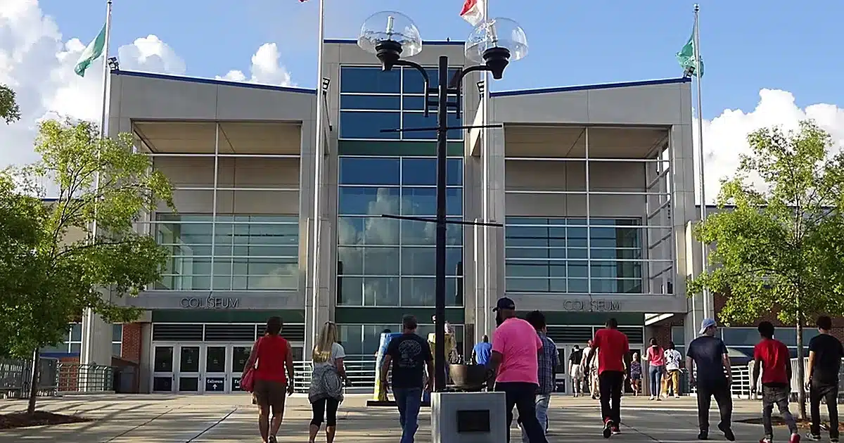 Front view of a modern glass Coliseum building with people walking toward the entrance on a sunny day.