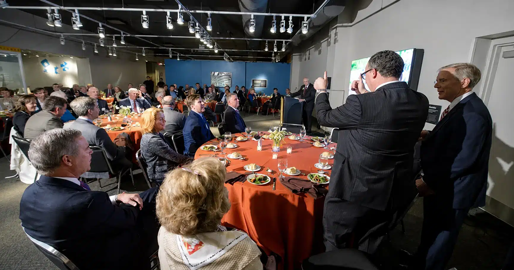 Large group of people in business attire seated around round tables with orange tablecloths, listening to a speaker at a podium.