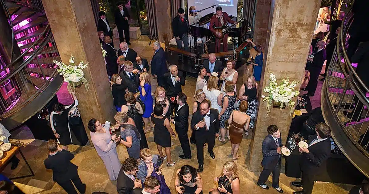 Crowded black-tie cocktail party in a grand hall, with a live band on a small stage and guests mingling.