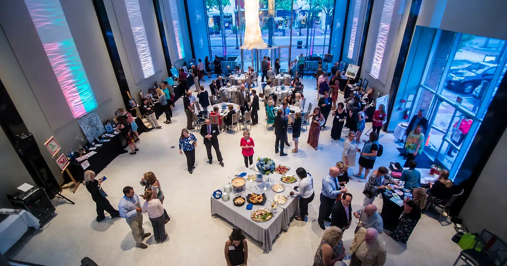 Wide atrium reception with guests mingling around a central buffet table in a bright modern lobby on display screens and blue lighting.