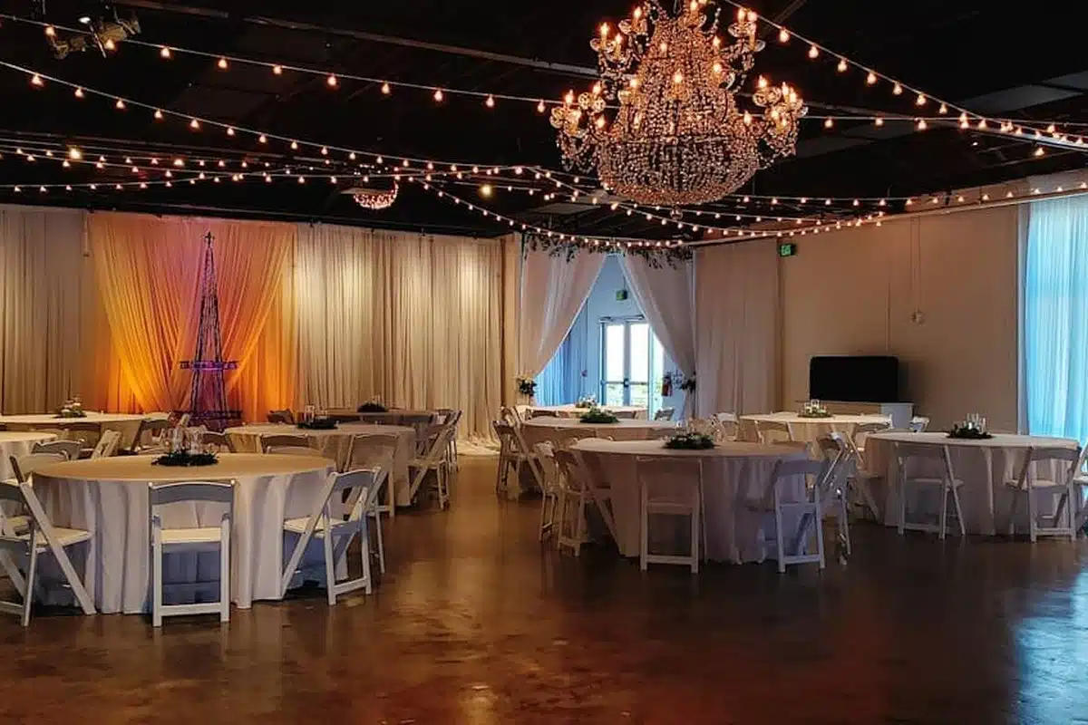 Elegant event hall with round tables in white linen, white chairs, draped curtains, and a crystal chandelier.