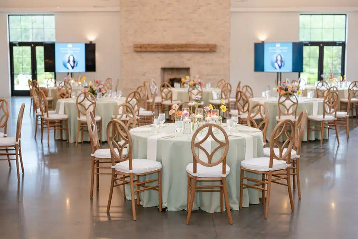 Banquet hall set for an event with round tables, pale green tablecloths, floral centerpieces, and wooden chairs with white cushions.
