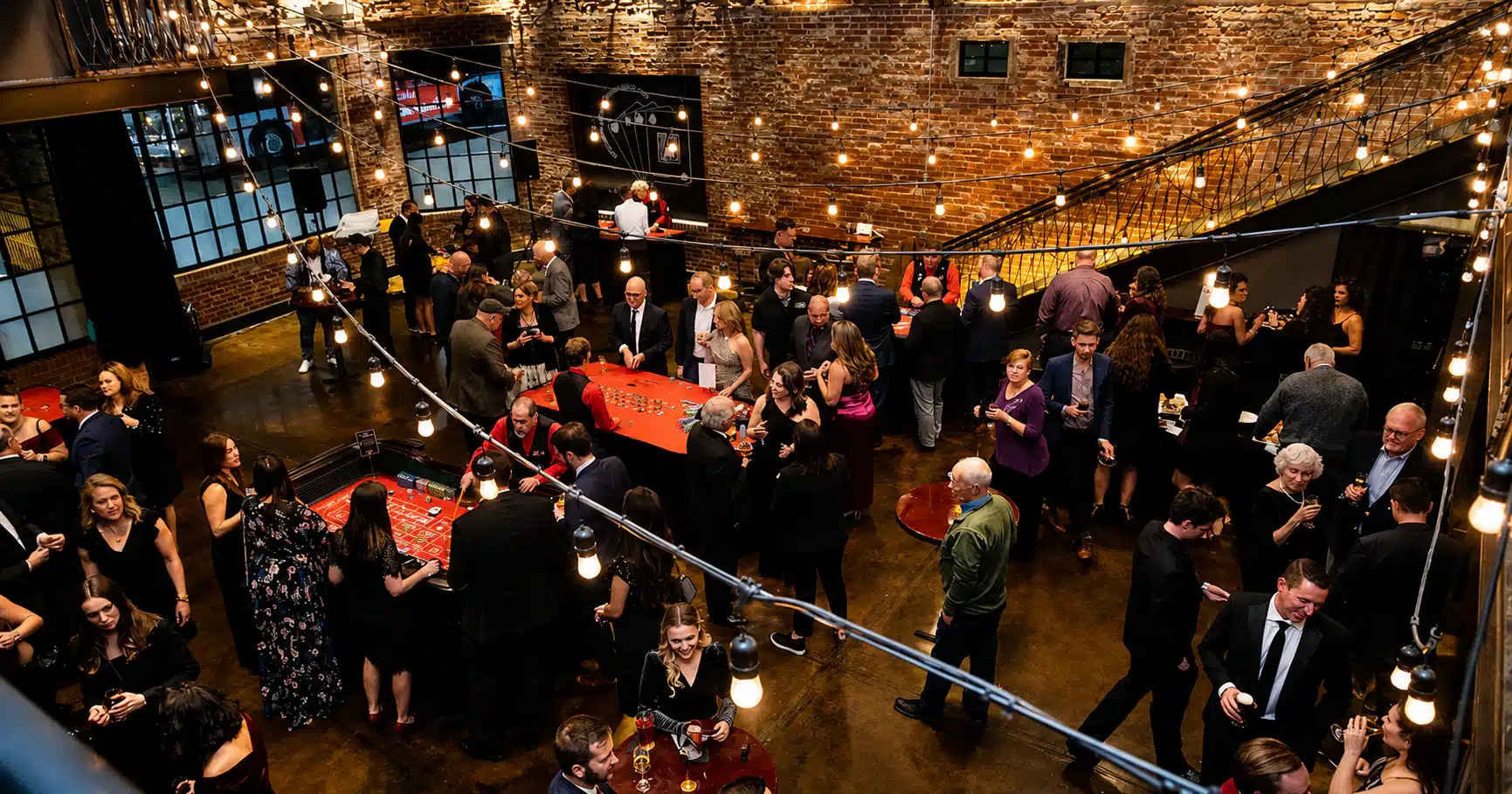 Crowd of well-dressed guests at a brick-walled venue with string lights, mingling around red casino tables at a social event.