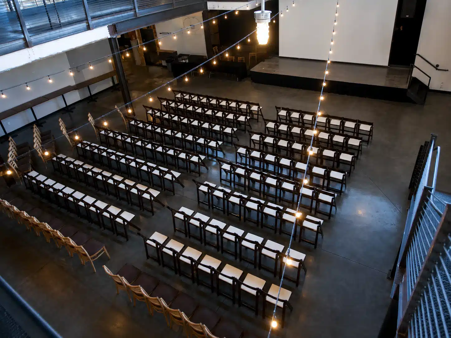 Industrial venue prepared for a presentation: rows of dark wooden chairs with white cushions face a small stage with a projector screen/backdrop.
