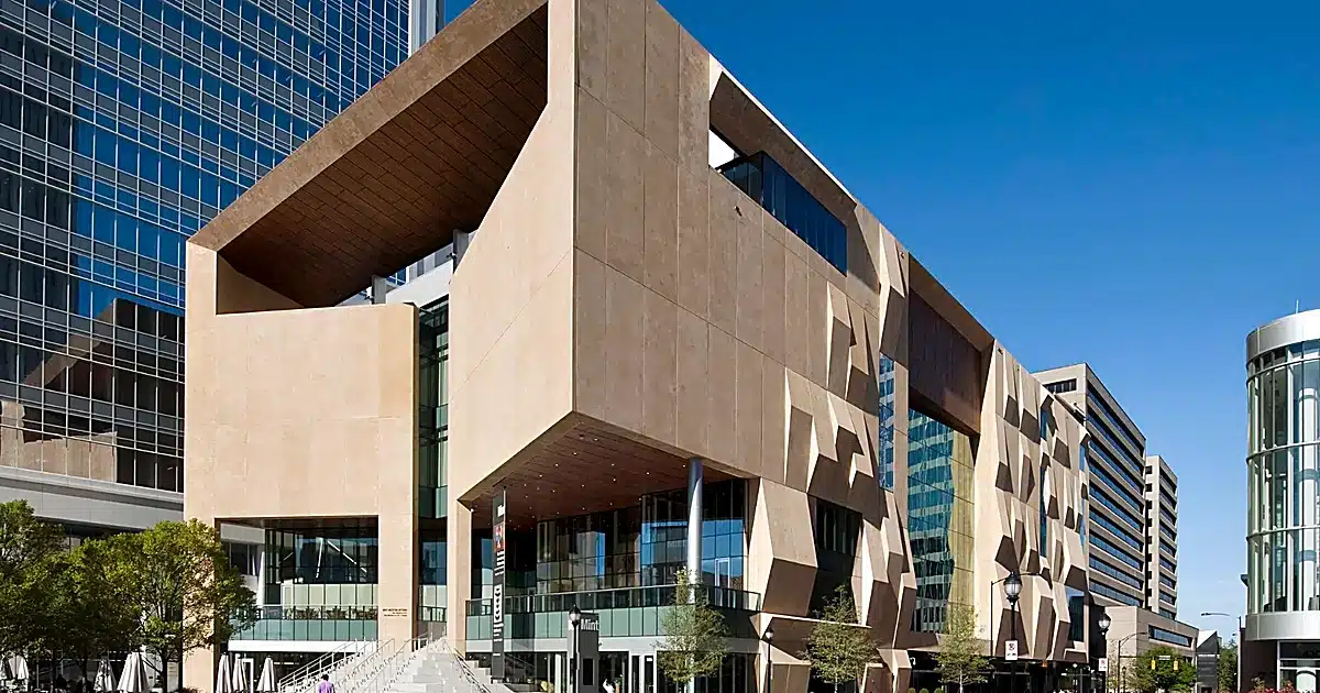 Modern beige geometric building with angular concrete blocks and glass façade under a clear blue sky.