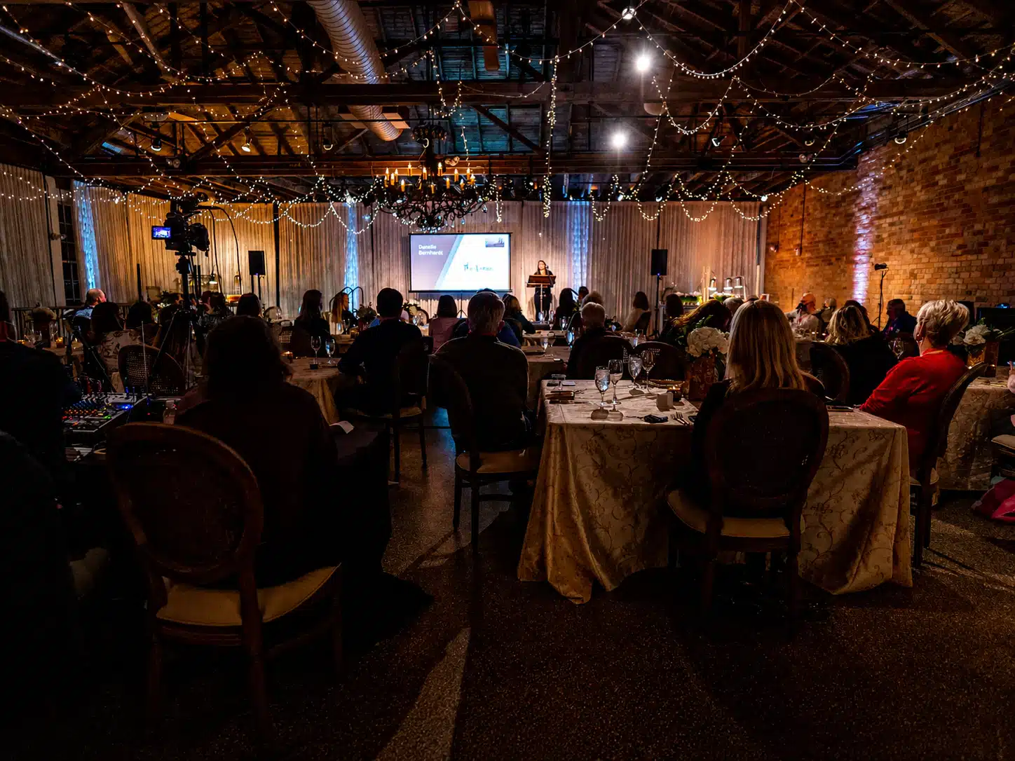 Formal banquet hall with string lights overhead and a stage; a speaker at a podium presents to seated guests at round-and-rectangle tables.
