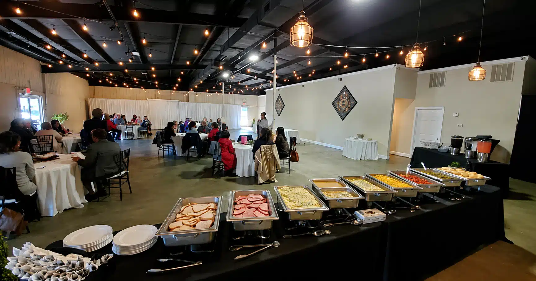 Banquet hall with guests seated at round tables while a long buffet with warming trays sits at the front under string lights.