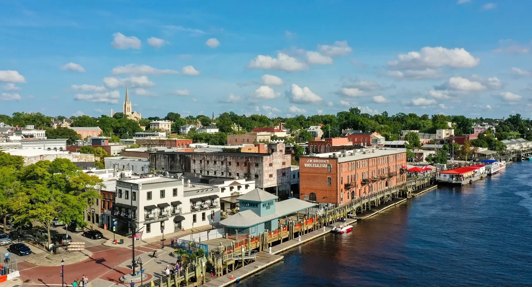 Aerial view of a riverside town with brick warehouses, a church steeple, and a boardwalk along the water.